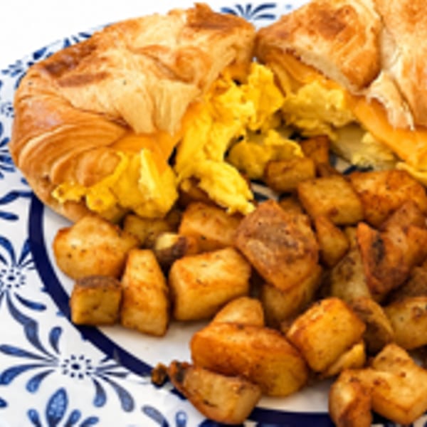 Croissant sandwich with scrambled eggs and cheese, served with seasoned breakfast potatoes on a patterned plate.