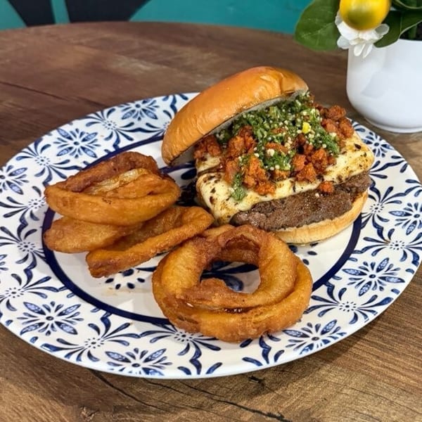 A burger with melted cheese, chorizo, and green relish, served with onion rings on a patterned plate.