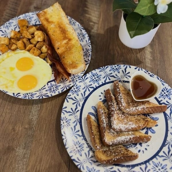 Two breakfast plates with eggs, bacon, potatoes, toast, and French toast with syrup on a wooden table.