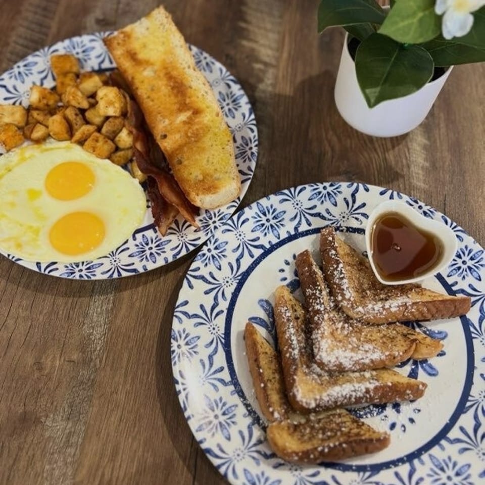 Two breakfast plates with eggs, bacon, potatoes, toast, and French toast with syrup on a wooden table.