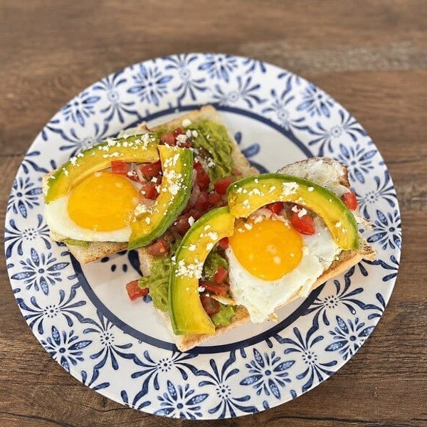 Two avocado toasts with fried eggs, diced tomatoes, and crumbled cheese on a patterned plate.