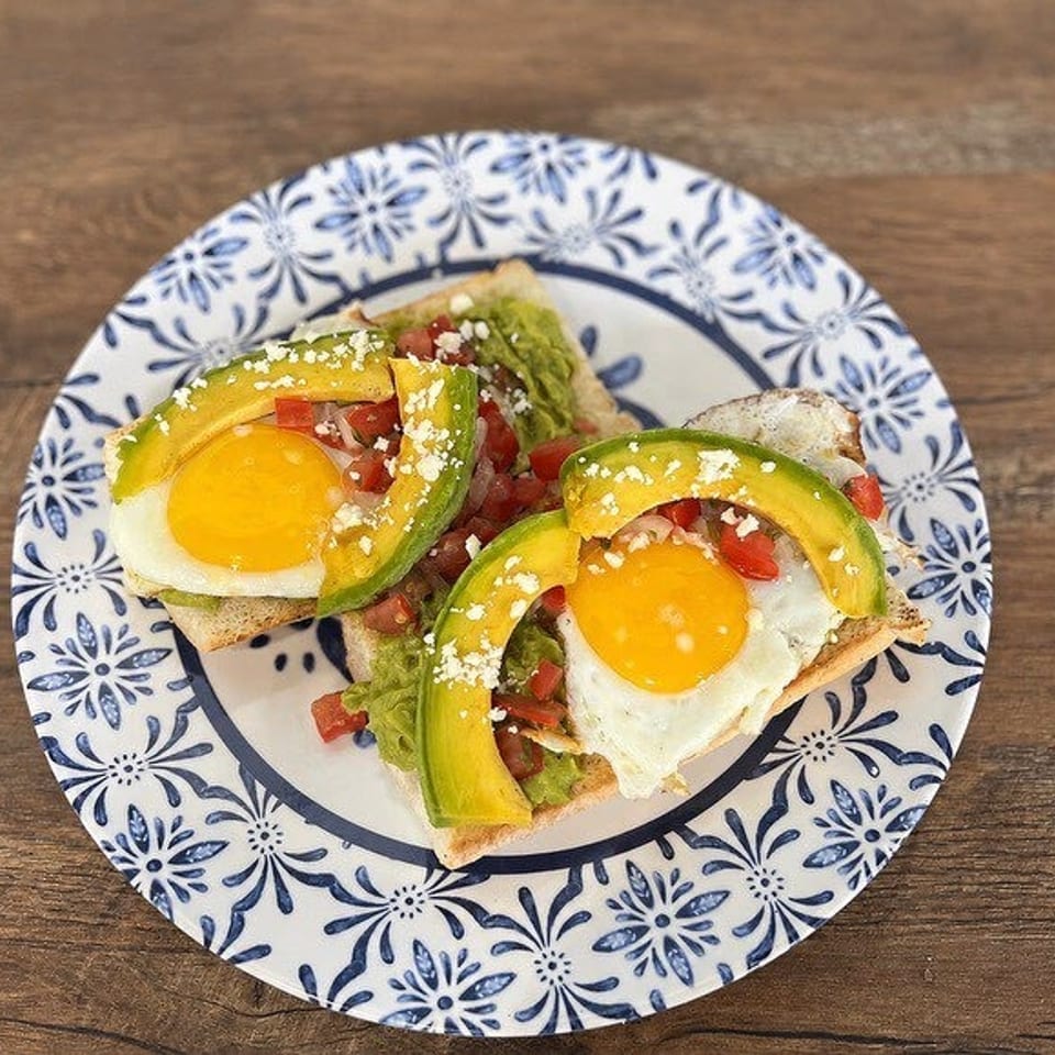 Two avocado toasts with fried eggs, diced tomatoes, and crumbled cheese on a patterned plate.