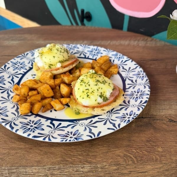 Eggs Benedict with ham and hollandaise sauce, served with crispy breakfast potatoes on a patterned plate.