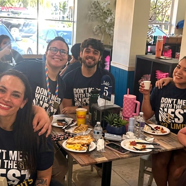 A group of smiling restaurant staff and patrons gathered around a table with food and drinks.