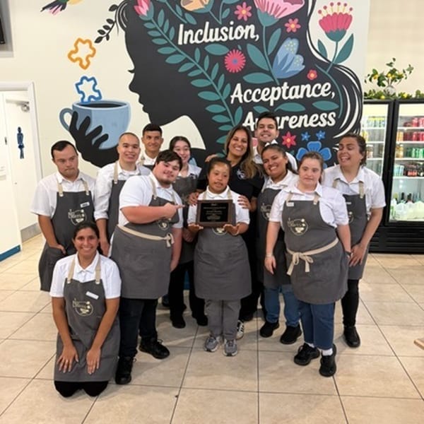 A group of diverse restaurant staff in aprons smiling, holding an award in front of a colorful mural.