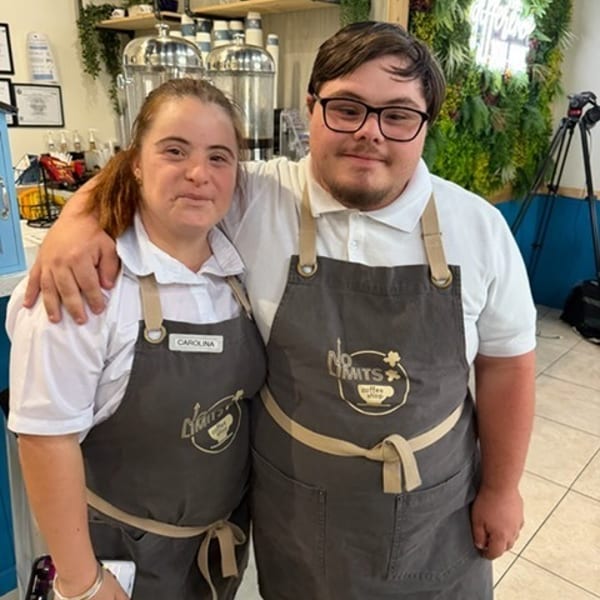 Two smiling employees in aprons, a woman named Carolina and a man, stand together in a coffee shop.